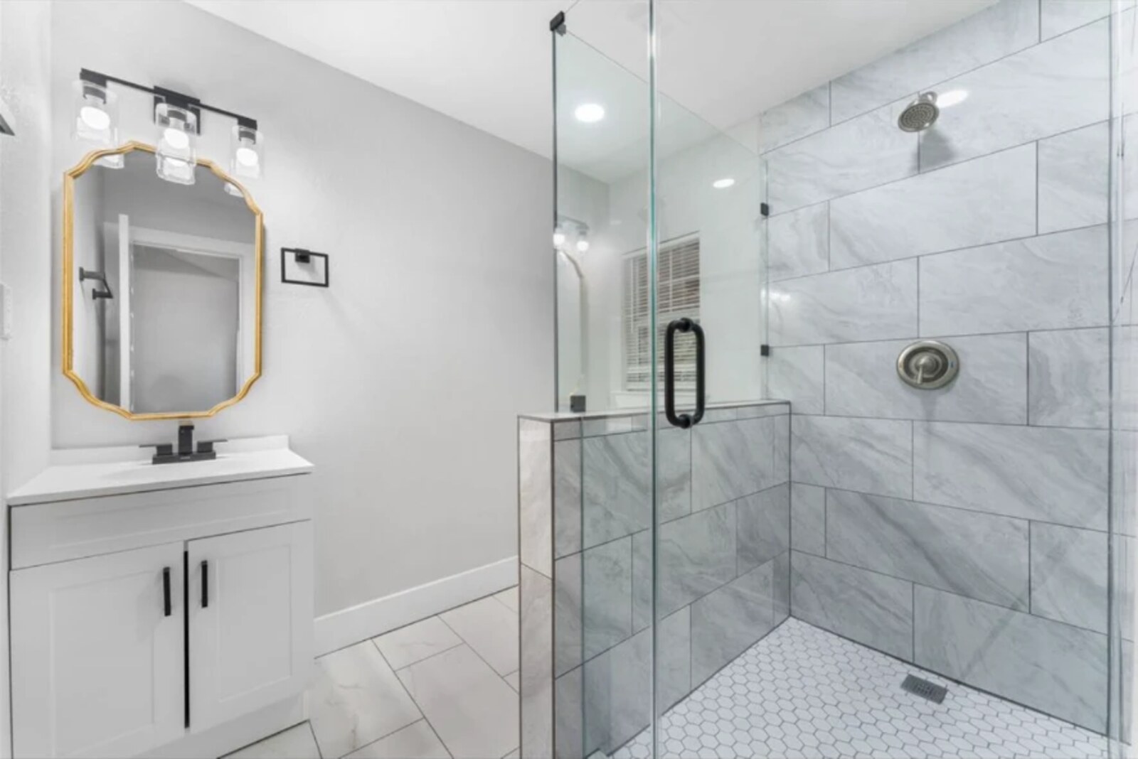Completed bathroom remodel with white vanity, gold-framed mirror, and walk-in glass shower with marble tile